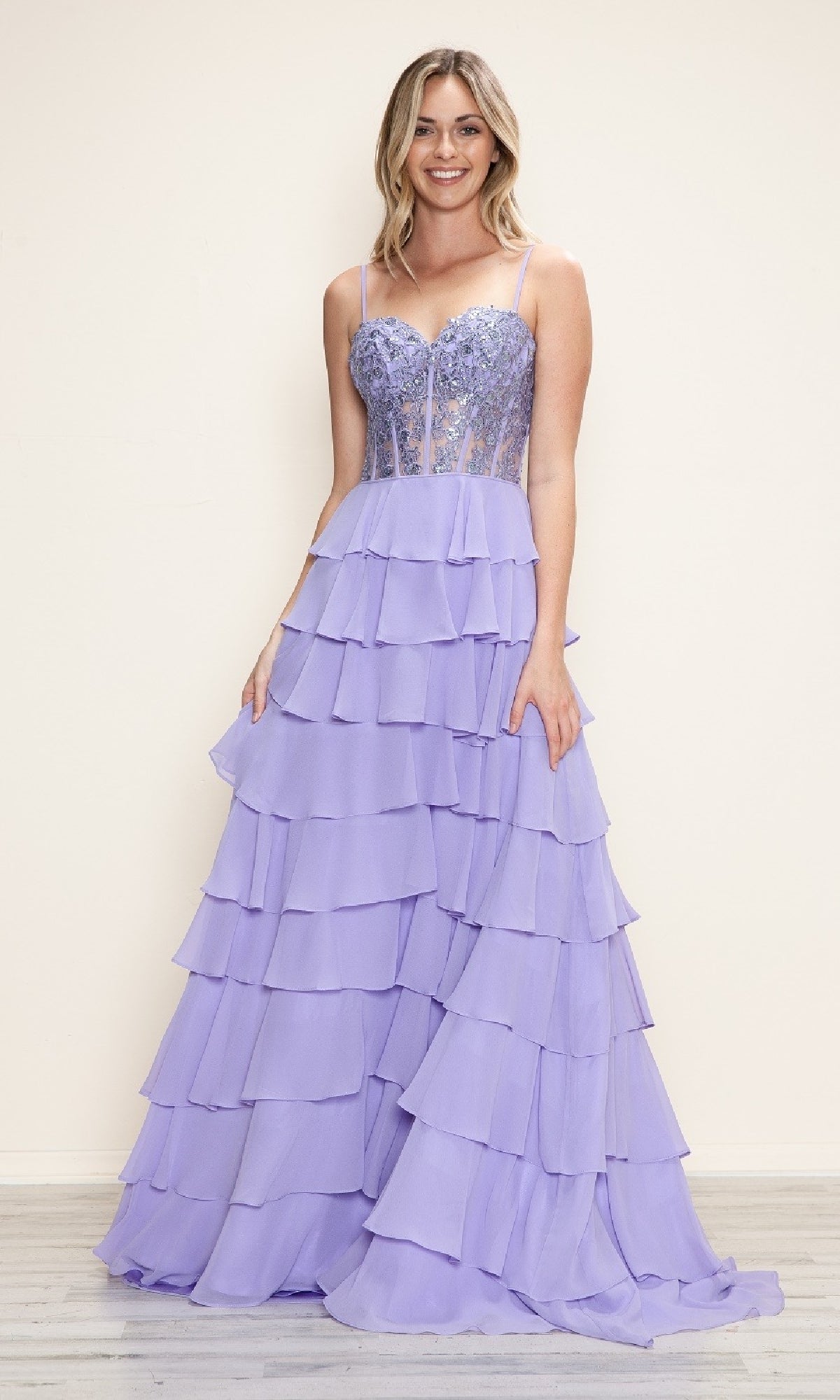 A woman stands smiling in the Long Tiered Corset Prom Dress: Poly USA 9698 in lavender, featuring a fitted corset-style bodice, thin straps, and a tiered a-line skirt, set against a simple light background.